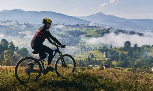 Man riding bicycle on grassy hill and looking at beautiful misty mountains. Male bicyclist enjoying panoramic view of majestic mountains during bicycle ride. Concept of sport, bicycling and nature.